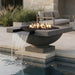 A modern outdoor fire pit bowl with a waterfall feature, set beside a swimming pool in a patio setting. The dark gray stone fire pit contains glowing orange and yellow fire rocks with active flames, and water cascades over a black ledge into the pool below.