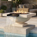 A modern outdoor fire pit bowl with a built-in waterfall feature, set on a stone patio above a swimming pool. The fire bowl contains dark river rocks and is lit with visible flames, with a light beige stone basin and dark metal spout.