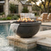 A round dark stone fire pit with active flames and black river rocks, featuring a waterfall spill into a swimming pool, set in a luxurious outdoor patio environment.