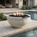 A round outdoor fire pit bowl with a waterfall spout, filled with black river rocks and active flames, situated beside a swimming pool on a stone patio.