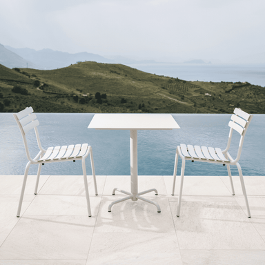 A white square outdoor coffee table and 2 white outdoor dining chairs placed beside the pool with view of a distant hills