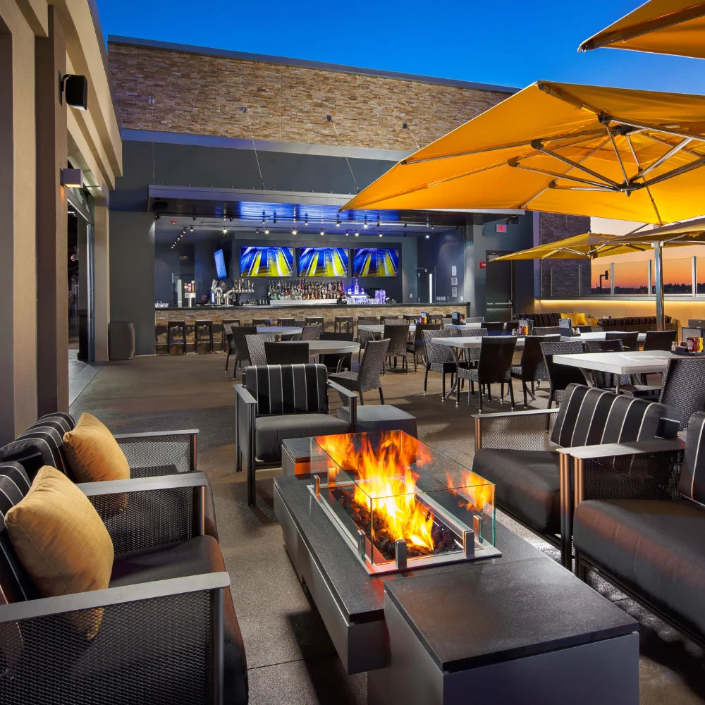 Modern outdoor restaurant seating with rectangular fire pit table, yellow umbrellas, and bar with multiple TVs at sunset.