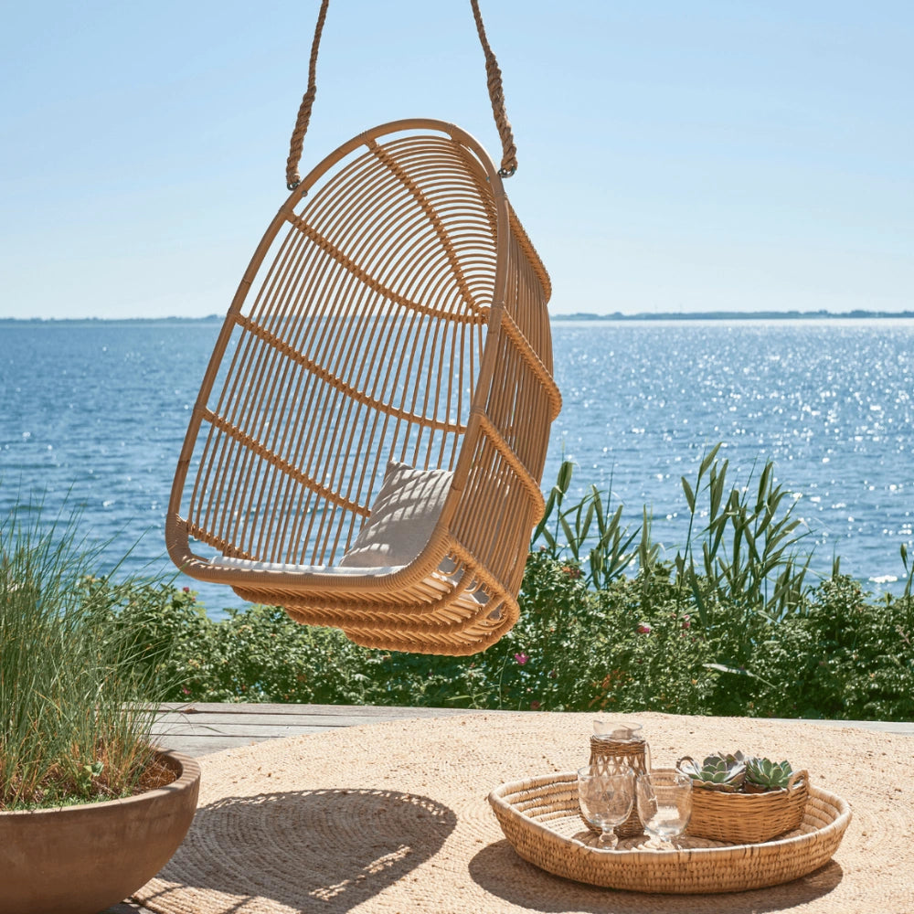 Natural rattan outdoor hanging swing chair beside a round serving tray on a seaside patio.