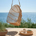 Natural rattan outdoor hanging swing chair beside a round serving tray on a seaside patio.