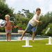 Two children playing outdoors by a swimming pool on a grassy lawn, using blue hoops and a green frisbee in a backyard setting with trees and a planter in the background.