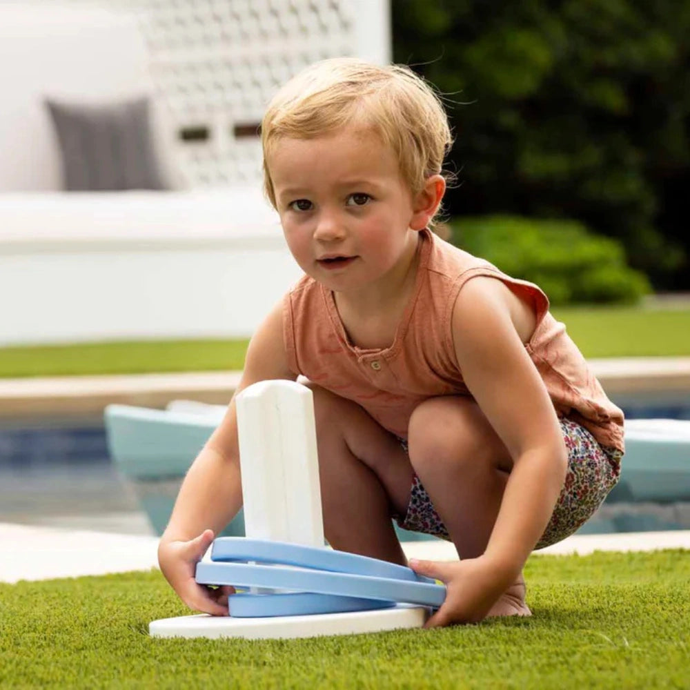 A young toddler with blonde hair crouches on a green lawn next to a swimming pool, holding a light blue and white pool toy.