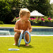 A toddler crouches on a green lawn by a swimming pool, holding light blue plastic rings. The background features a swimming pool, lush green trees, and a patio umbrella.