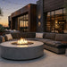 A modern outdoor patio setup at dusk featuring a round concrete fire pit with active flames and a matching curved sectional sofa in dark gray cushions with white pillows, set against a luxury resort-style building with warm lit windows.