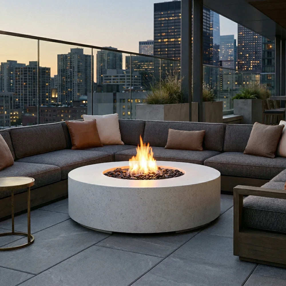 A round concrete fire pit with a glowing flame sits in the center of a modern outdoor sectional sofa on a rooftop terrace at dusk, with a city skyline in the background.