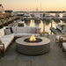 Outdoor patio furniture set at a marina at sunset, featuring a round concrete fire pit with a visible flame, a sectional sofa with light beige cushions, and matching armchairs on a stone tile deck with boats in the background.
