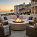 Outdoor patio furniture set with a round concrete fire pit, curved sectional sofa, and two wicker armchairs on a stone patio in front of a large coastal mansion at sunset.