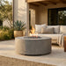 A round concrete fire pit table with a lit flame, set on a stone patio in front of a modern outdoor sofa.