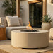 A round beige outdoor fire pit table with a visible flame and black fire rocks, placed on a patio beside a light beige sofa in a modern patio setting.