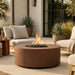 Round outdoor fire pit table with a rusted metal finish, shown lit on a patio with beige lounge chairs, potted plants, and a modern building facade in the background.