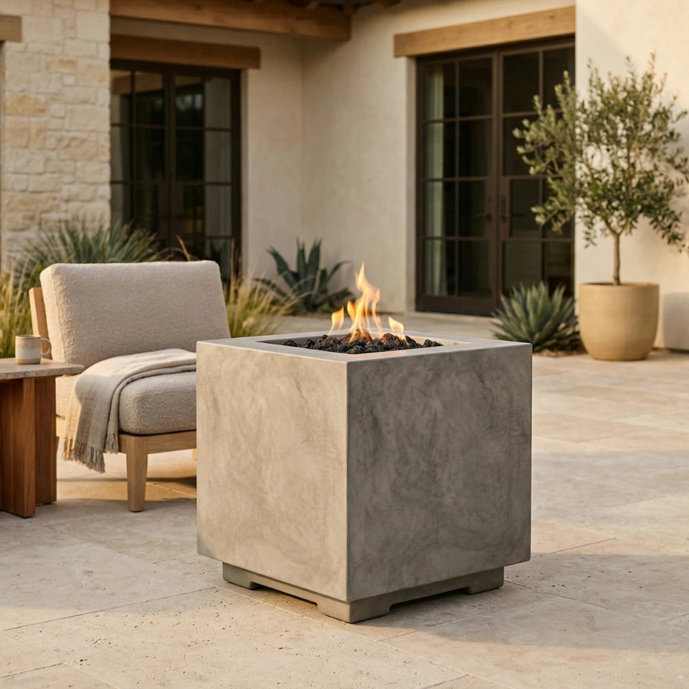 A square concrete fire pit table with a visible flame, placed on a stone patio in front of a modern house. A light beige armchair and potted plants are visible in the background.