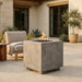 A square concrete fire pit table with a visible flame, placed on a stone patio in front of a modern house. A light beige armchair and potted plants are visible in the background.