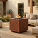 A square outdoor fire pit with a copper-toned metal exterior and active flames, placed on a patio next to a light beige sofa in a modern outdoor living space.