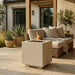 A square concrete fire pit table with a lit flame sits on a stone patio in front of a wicker sectional sofa with neutral cushions and a throw blanket. The setting includes potted plants and a house exterior.