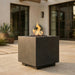A square concrete fire pit with a dark gray finish, featuring a flame burning over black rocks inside, set on a stone patio in front of a modern house.