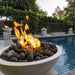 A four white round concrete fire pit bowl filled with lava rocks with flames placed on a concrete pedestal beside the pool
