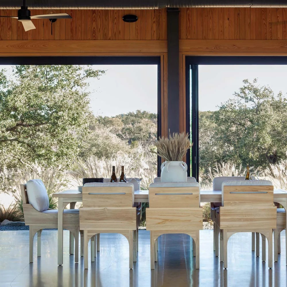 Dining area with wooden outdoor dining table and chairs in front of large glass doors with a view of trees.