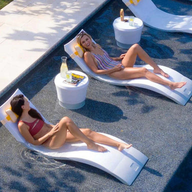 Two women lounging on white in-pool chaise loungers with side tables on a pool ledge.