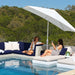 Outdoor poolside lounge scene with two women sitting on a modern sectional sofa and floating daybeds under a large white patio umbrella, with a forested hillside in the background.