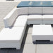White outdoor sectional sofa with a matching square coffee table on a light tiled patio. The sectional has a U-shaped arrangement with thick cushioned seat and back cushions, and a blue patterned rectangular rug or mat is visible in the background.