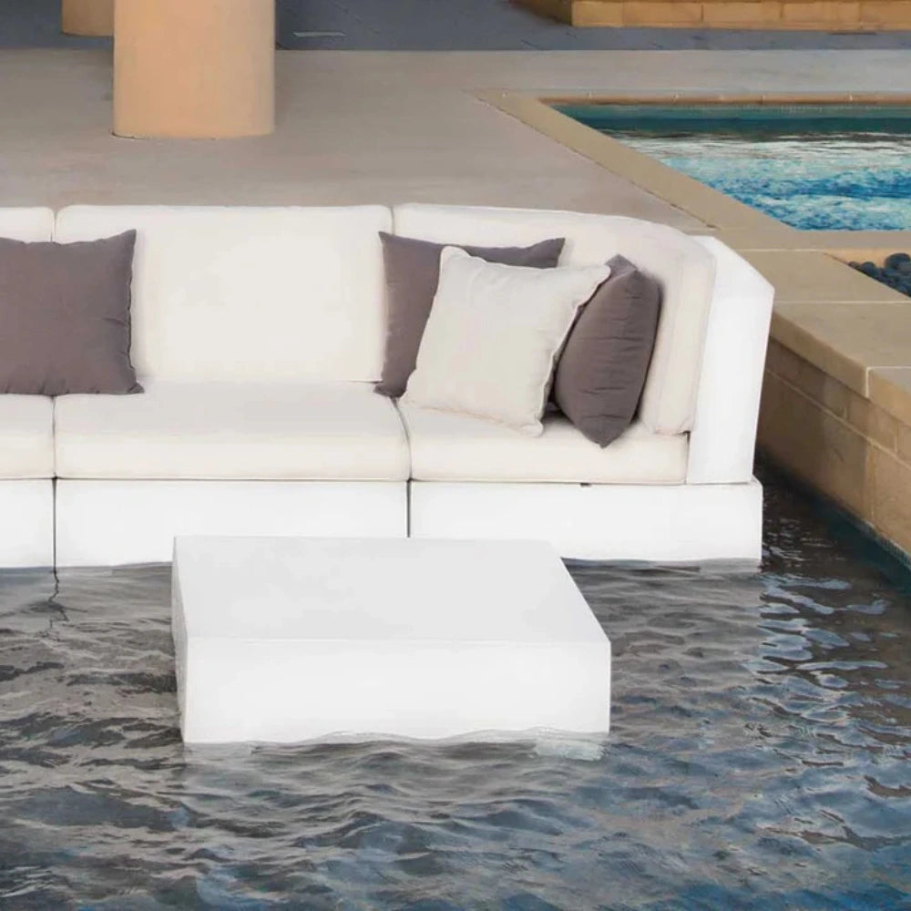 A modern outdoor sectional sofa in off-white with dark gray and light beige accent pillows, positioned next to a swimming pool with a matching white rectangular coffee table in the foreground.