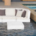 A modern outdoor sectional sofa in off-white with dark gray and light beige accent pillows, positioned next to a swimming pool with a matching white rectangular coffee table in the foreground.