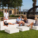 A white outdoor sectional sofa set with matching cube coffee tables, shown on a grassy patio with several people sitting and standing around it. Trees, a large hanging chair, and buildings are visible in the background.
