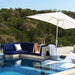 Outdoor in-pool lounge scene with a woman reclining on a navy and gray sectional sofa under a large white patio umbrella. The setting includes a swimming pool, deck, and dense green trees in the background.