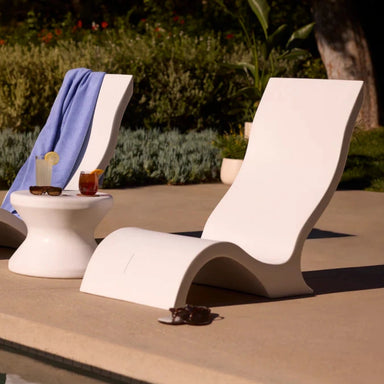 Two modern white tanning ledge chairs with matching side table on a sunlit poolside, surrounded by greenery.