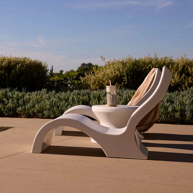 Side view of a pair of white in-pool lounge chairs with a matching side table in a landscaped pool area.