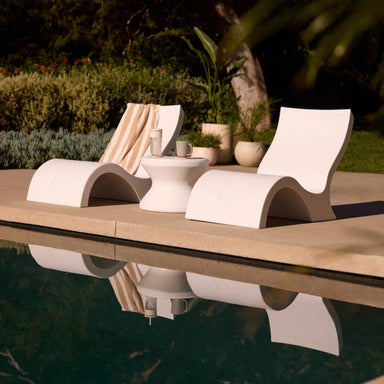 Two modern white tanning ledge chairs with a matching side table beside a pool, surrounded by greenery.
