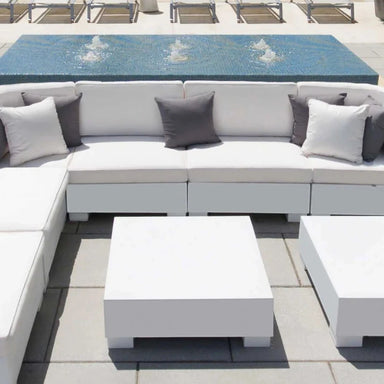 White outdoor sectional sofa with gray accent pillows, matching white coffee tables, and a blue tiled pool deck in the background.