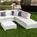 White outdoor sectional sofa set with a matching ottoman on a green lawn, featuring cream cushions, gray accent pillows, and a modern patio setting with a wicker chair in the background.