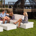 Outdoor patio scene with three people sitting on a white modular sectional sofa and cube tables on green grass, with a large black woven pod structure and city buildings in the background.
