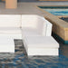 White modular outdoor poolside seating with thick cushions, shown partially submerged in blue water next to a beige stone deck and pool edge.