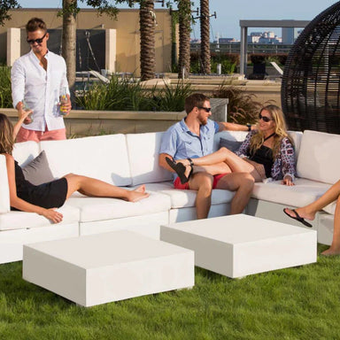 Outdoor patio with a white sectional sofa and coffee tables , with people sitting and standing around it. 