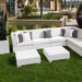 White outdoor in-pool sectional sofa set with gray and white throw pillows, two matching white cube in-pool coffee tables, and a green lawn in front of a resort-style patio background.