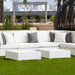 A modern outdoor in-pool sectional sofa set in bright white with gray throw pillows, paired with two matching white cube in-pool coffee tables, set on a green lawn with a city skyline and patio furniture in the background.