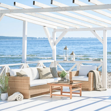Natural rattan outdoor lounge chair with white cushions beside a matching 3-seater sofa and teak coffee table.