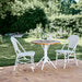 White rattan outdoor dining side chair beside a round teak café table in a garden setting.