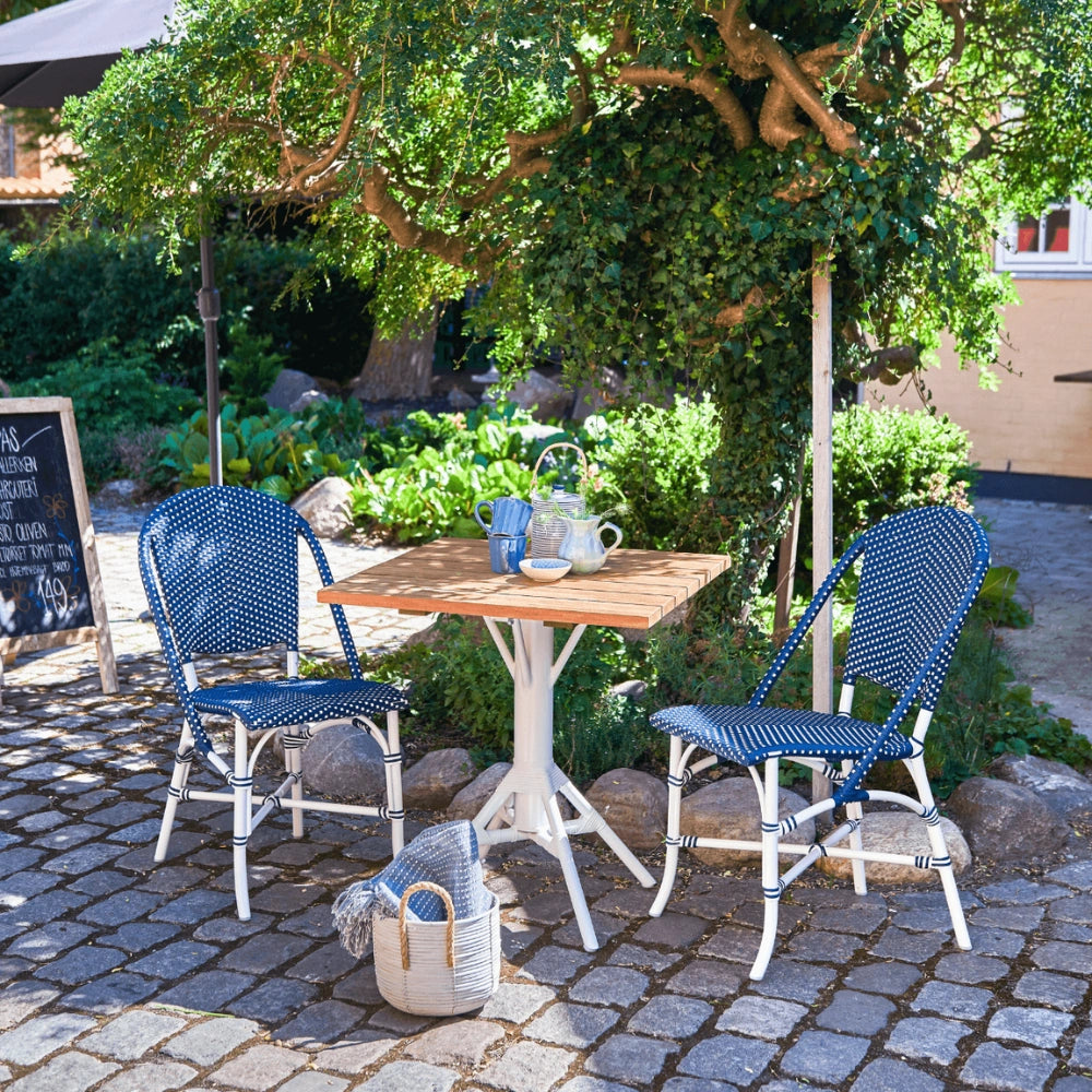 Blue and white rattan outdoor dining side chair beside a square teak café table in a garden setting.