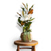 Potted magnolia tree plant with white blooms and green leaves, wrapped in burlap and tied with a green ribbon, placed on a wooden stool.