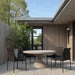 Modern beige round concrete outdoor dining table, black wicker chairs and greenery on a wooden deck patio