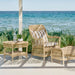 Natural rattan outdoor square side table beside a matching lounge chair and footstool on a seaside patio.