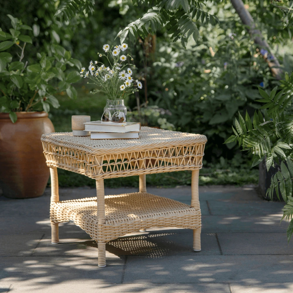 Natural rattan outdoor square side table with a woven lower shelf on a garden patio.