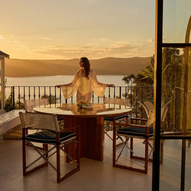 Balcony with round wooden dining table and chairs woman in light robe leaning on railing overlooking sea and distant hills.